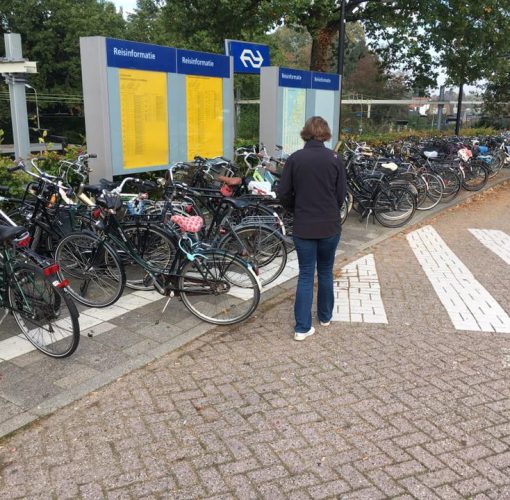 Foto van een vrouw die noodgedwongen op de straat loopt, de geleidelijnen op de stoep zijn geblokkeerd door fietsen. Er zijn vertrekborden van de trein zichtbaar.